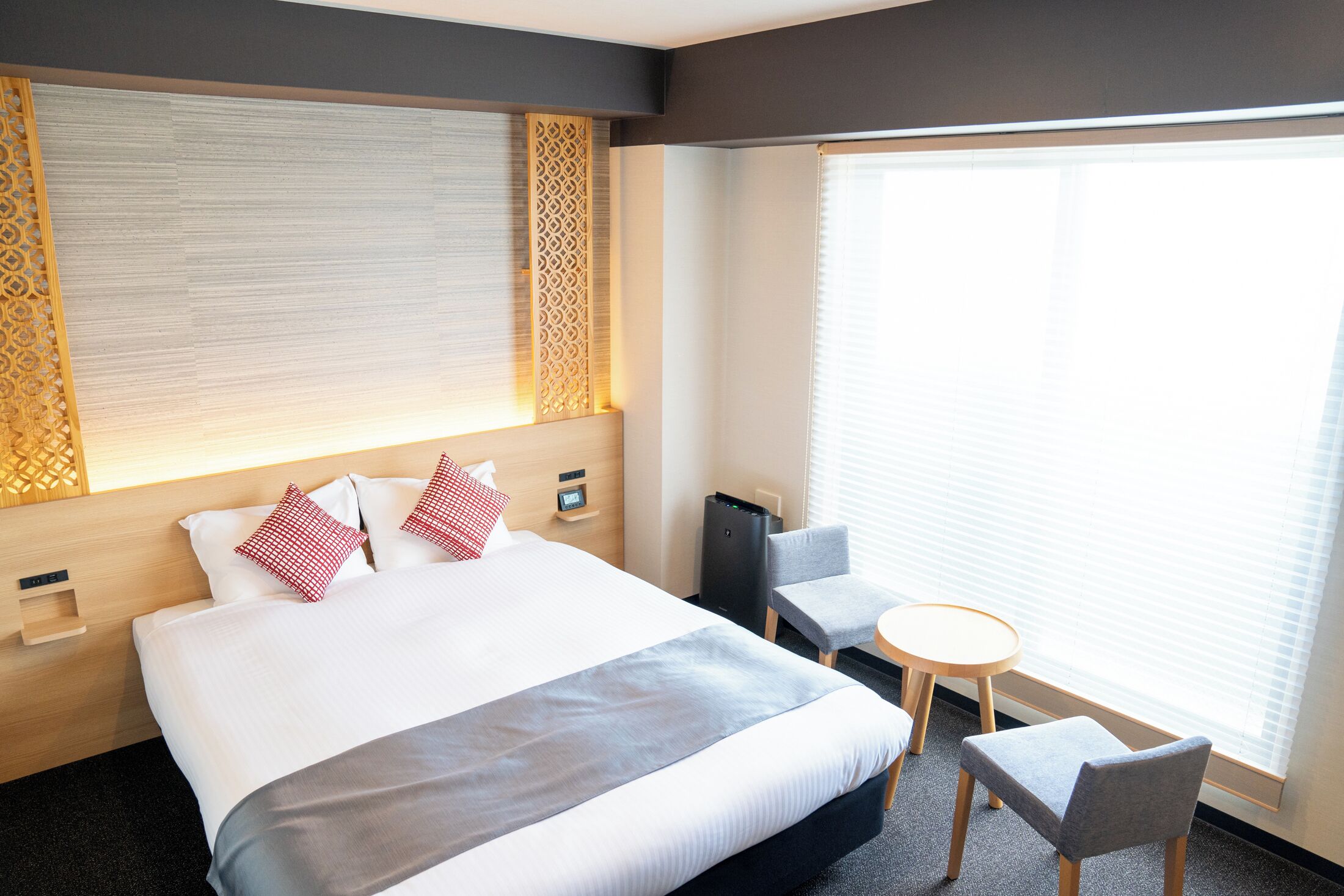 Ein Hotelzimmer mit einem großen Bett, einem Nachttisch, einem Sessel, einem kleinen Tisch und einem Fenster mit Jalousien.