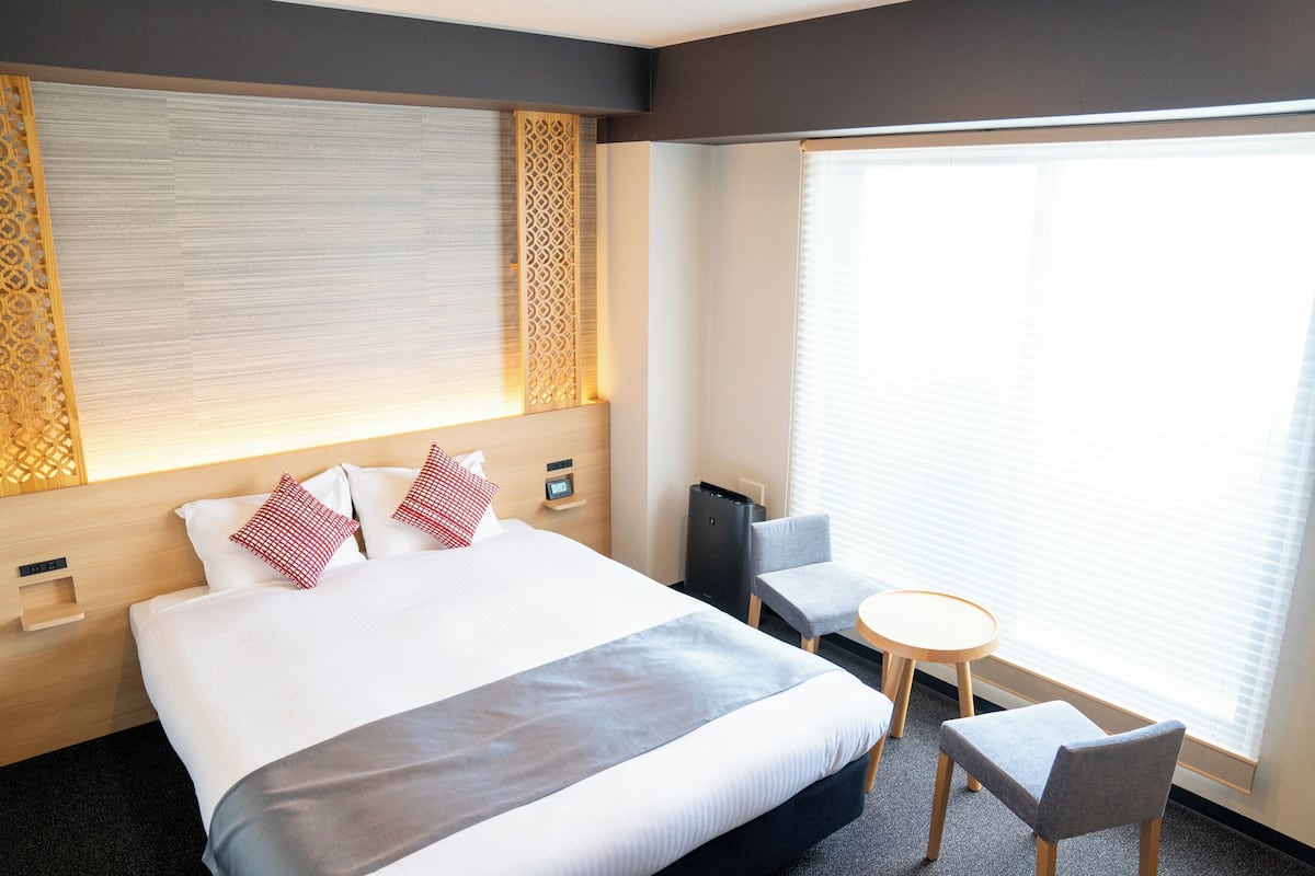 A hotel room with a large bed, a bedside table, a chair, a small table, and a window with blinds.