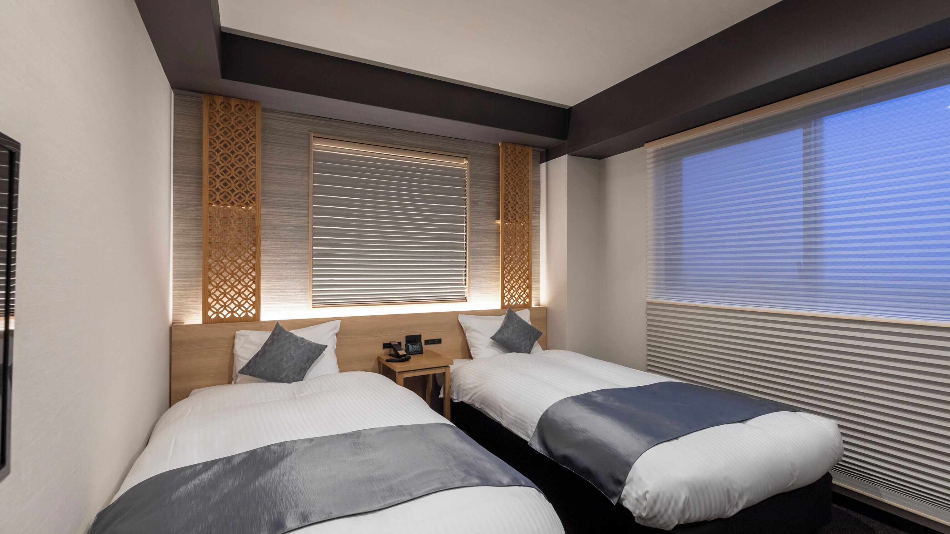 Une chambre d’hôtel avec deux lits, une tête de lit en bois, une table de chevet et une fenêtre avec des stores.