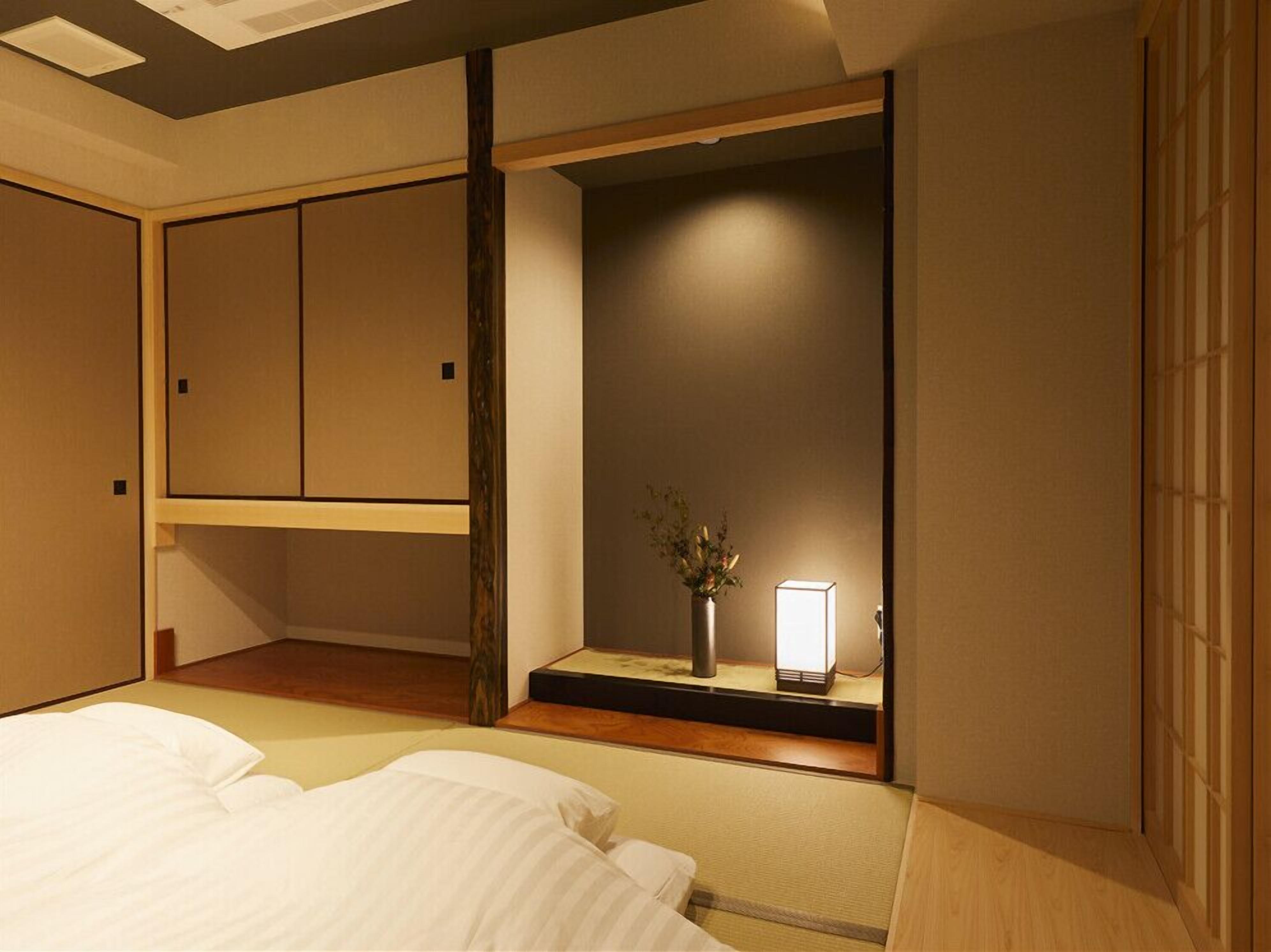Habitación de hotel con cama, mesita de noche con lámpara, pequeño florero con flores y armario con puerta corredera.