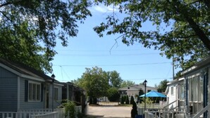 A narrow street flanked by mobile homes with white railings, a clear sky, and a blue umbrella in the distance. - Cozy 2BR Cottage • Walk to Beach 1 • Free Parking & WiFi (Wasaga Beach)