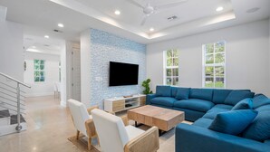 Uma sala de estar moderna com um sofá azul em forma de L, cadeiras brancas, uma mesa de centro de madeira e uma TV de tela plana fixada em uma parede com textura de tijolos.
