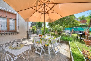Een terras met een tafel onder een grote parasol, omringd door witte stoelen, en een witte wagen met een dienblad.