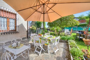 Een terras buiten met een eettafel, omringd door witte stoelen, onder een grote parasol.