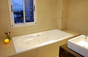 Une salle de bain moderne avec une grande baignoire, une fenêtre donnant sur des immeubles et un petit vase avec des fleurs.