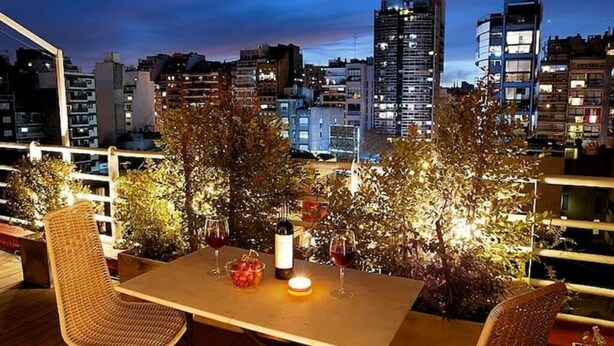 En tagterrasse med et bord dækket til to, omgivet af krukker med planter og udsigt over byens skyline i skumringen.