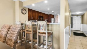 A dining area with wicker chairs, a wooden table, and a kitchen in the background. - Destin Towers 32 (Destin)