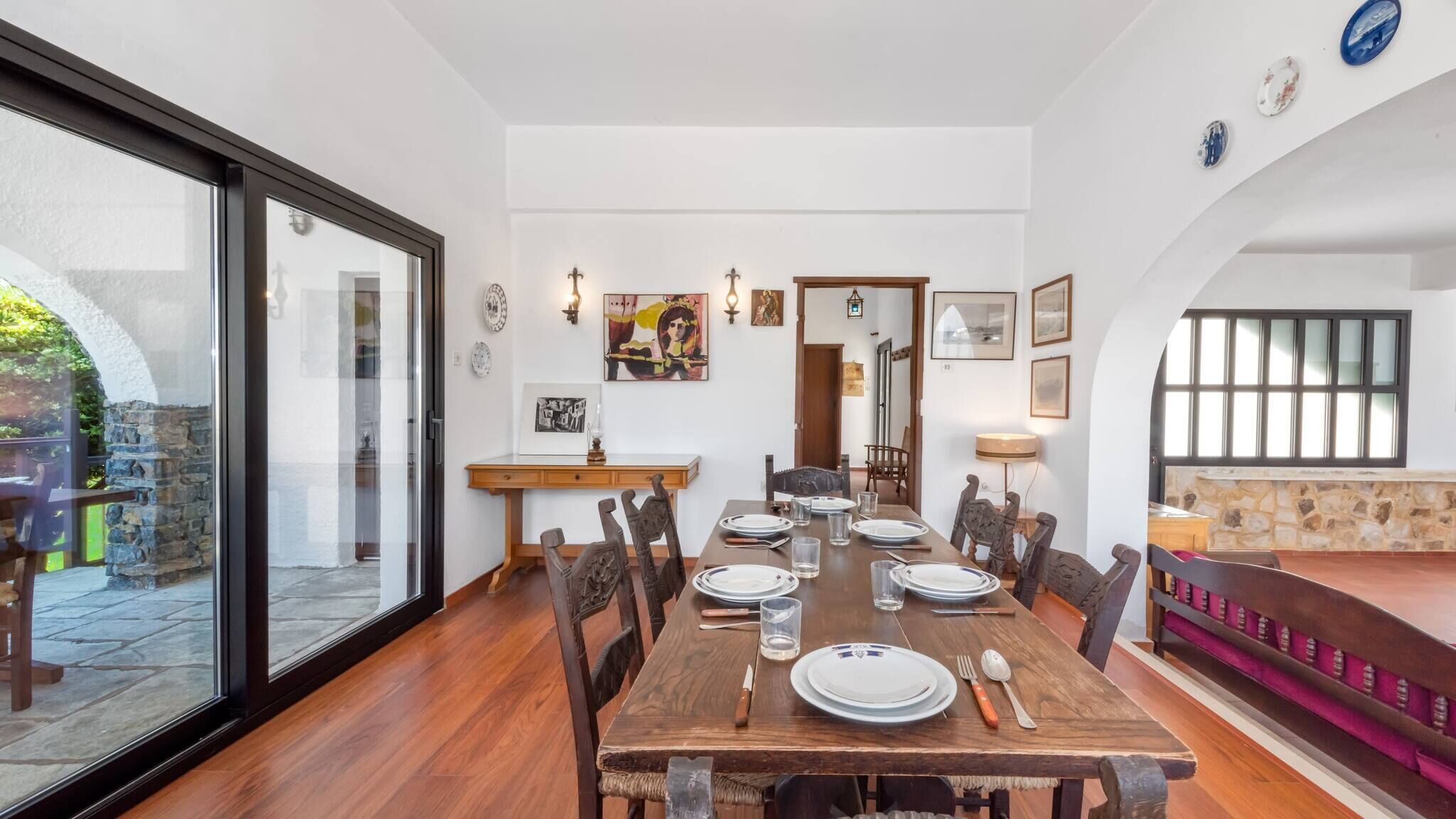 A dining area with a wooden table set for six, surrounded by chairs, with a stone wall and a glass door leading outside.