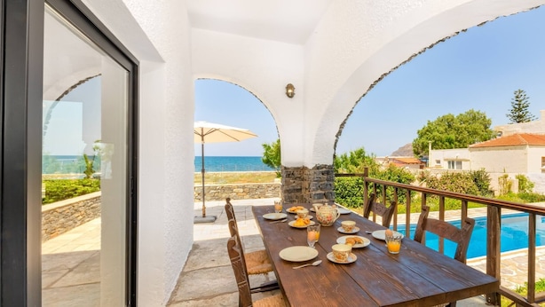 A wooden dining table with plates, glasses, and a fruit bowl set outdoors with a view of the sea.
