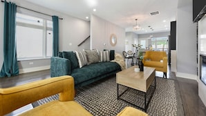 En moderne stue med en mørkegrøn sofa, en gul lænestol og et sofabord. I baggrunden ses et spiseområde med et rundt bord og stole.