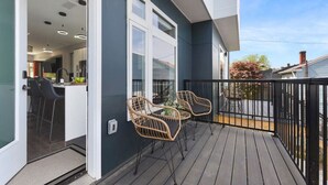 A balcony with two wicker chairs and a small table, a sliding glass door leading to a kitchen area, and a view of neighboring houses. - ★ HostWise Stays ★ Rooftop Deck & Fire Pit Haven | Theater & Balcony (Pittsburgh)