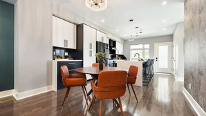 A modern kitchen with a central island, wooden flooring, and a dining area with orange chairs. - ★ HostWise Stays ★ Rooftop Deck & Fire Pit Haven | Theater & Balcony (Pittsburgh)