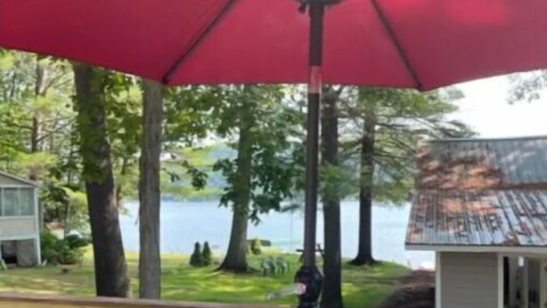 A deck with a round glass table and chairs, a red umbrella, and a view of a lake and trees. - Lake St Catherine Cabin For Two with deck and lake access (Poultney)
