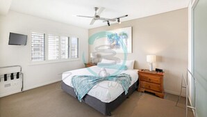 Ein Schlafzimmer mit einem Bett, einem Deckenventilator, einem Nachttisch mit Lampe, einem an der Wand befestigten Fernseher und einem Fenster mit Jalousien.
