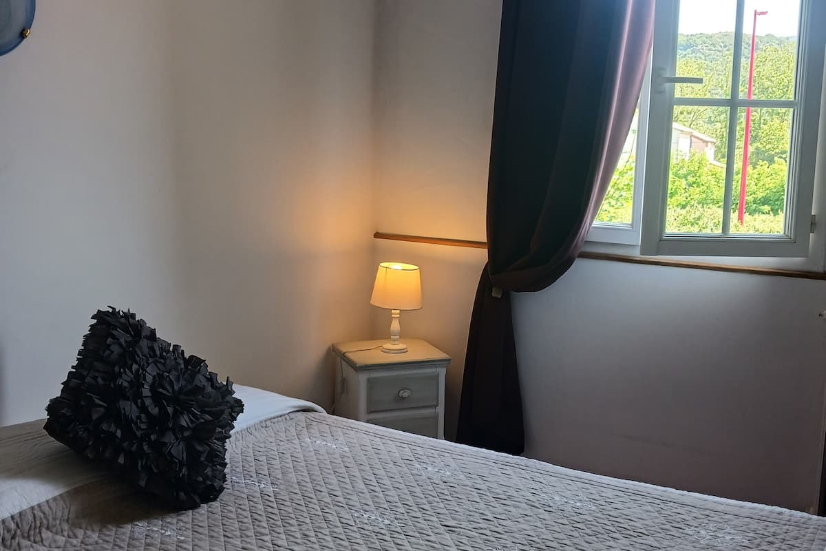 Une chambre à coucher avec un lit, une table de chevet avec une lampe, une fenêtre avec des rideaux et une vue sur la verdure à l’extérieur.