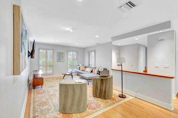 Un salon moderne avec un canapé, une table basse et des objets de décoration. De grandes fenêtres sont équipées de stores, et on y trouve également une lampe sur pied et un plancher en bois.