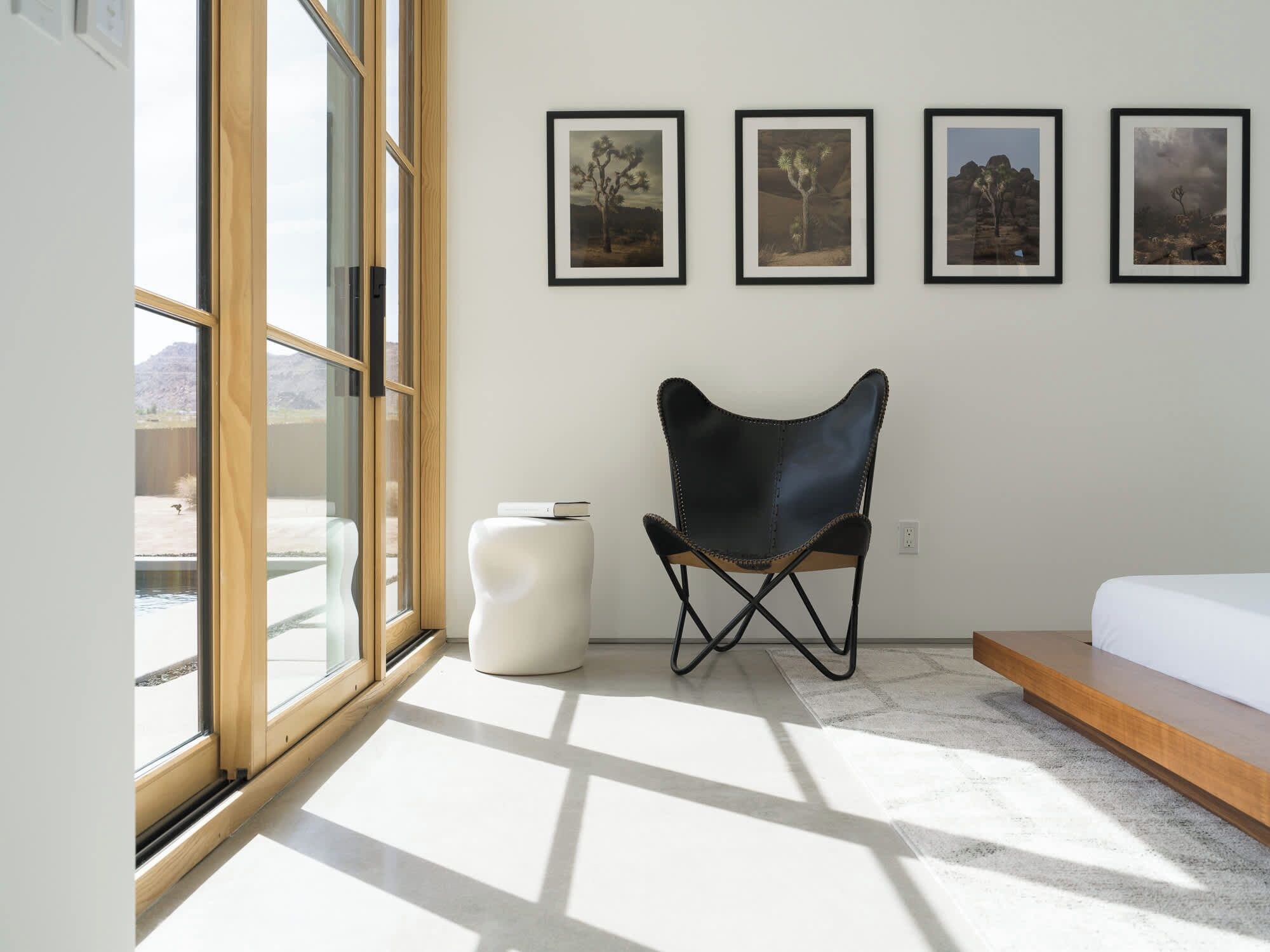 Un dormitorio moderno con un amplio ventanal, un sillón de cuero negro, un reposapiés blanco y una cama con cabecero de madera.