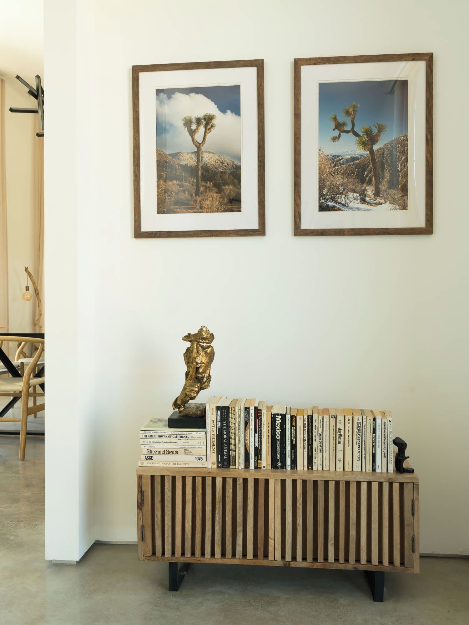 Una estantería de madera con libros y una estatua dorada, dos cuadros en la pared, y una silla al fondo.