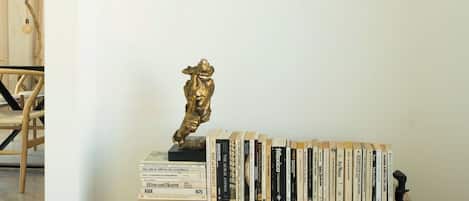Una estantería de madera con libros y una estatua dorada, dos cuadros en la pared, y una silla al fondo.