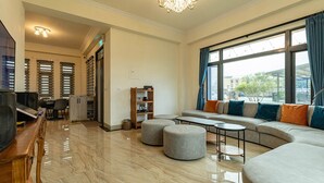 Amplia sala de estar con un sofá grande, otomanas, una mesa de centro y un televisor. Hay una estantería y un escritorio con una silla. La habitación tiene ventanas grandes con cortinas.