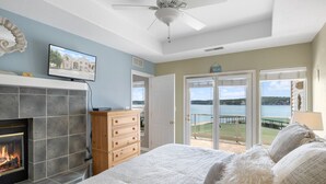 A bedroom with a fireplace, a television, a bed with white bedding, a wooden dresser, and a view of a lake through a glass door. - Ozark Overlook – Waterfront, Two Pools, Tennis, and More! (Lake Ozark)