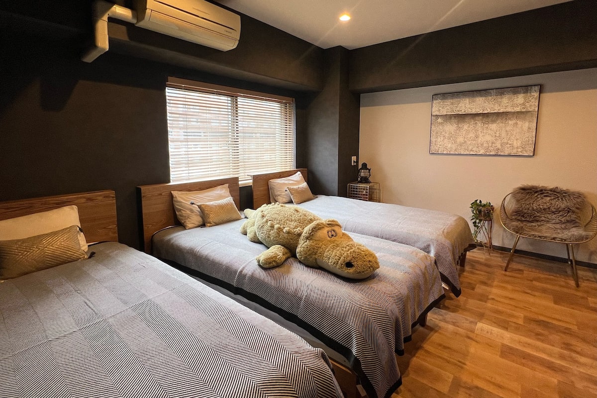 A hotel room with two beds, a large teddy bear on one bed, a chair, and a wall-mounted air conditioner.