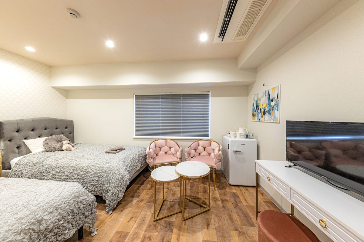 A bedroom with two beds, a TV, a small fridge, and a window with blinds.