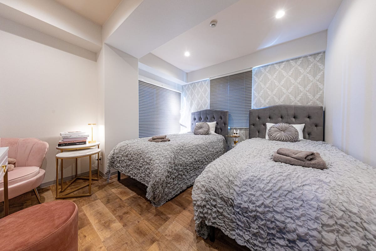 A bedroom with two beds, a small table, a chair, and a window with blinds.