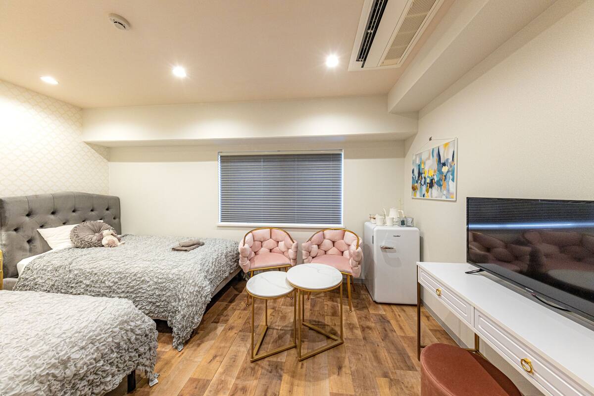 A bedroom with two beds, a TV, a small fridge, and a window with blinds.