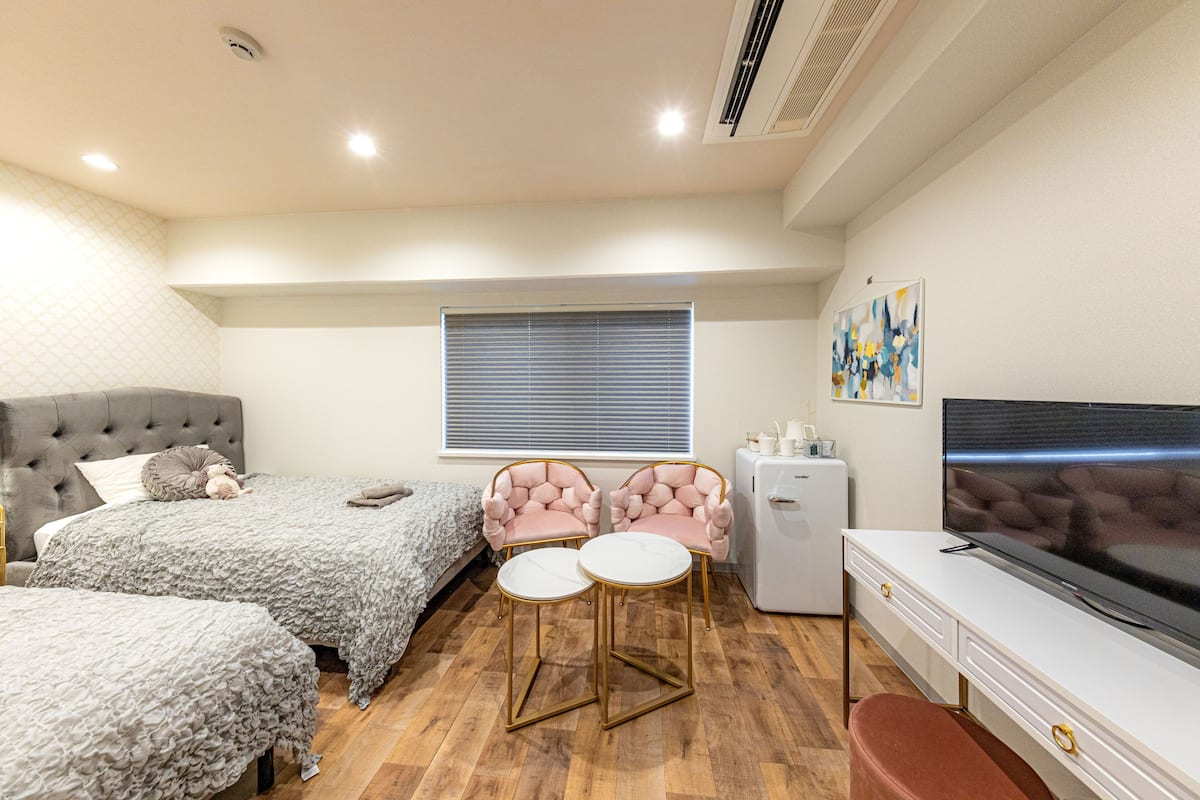 A bedroom with two beds, a TV, a small fridge, and a window with blinds.