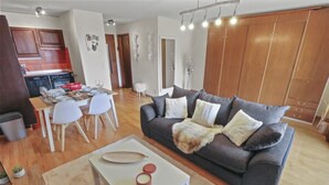 Un salon moderne avec un canapé gris, des chaises blanches, une table à manger dressée pour quatre personnes et une cuisine en arrière-plan.