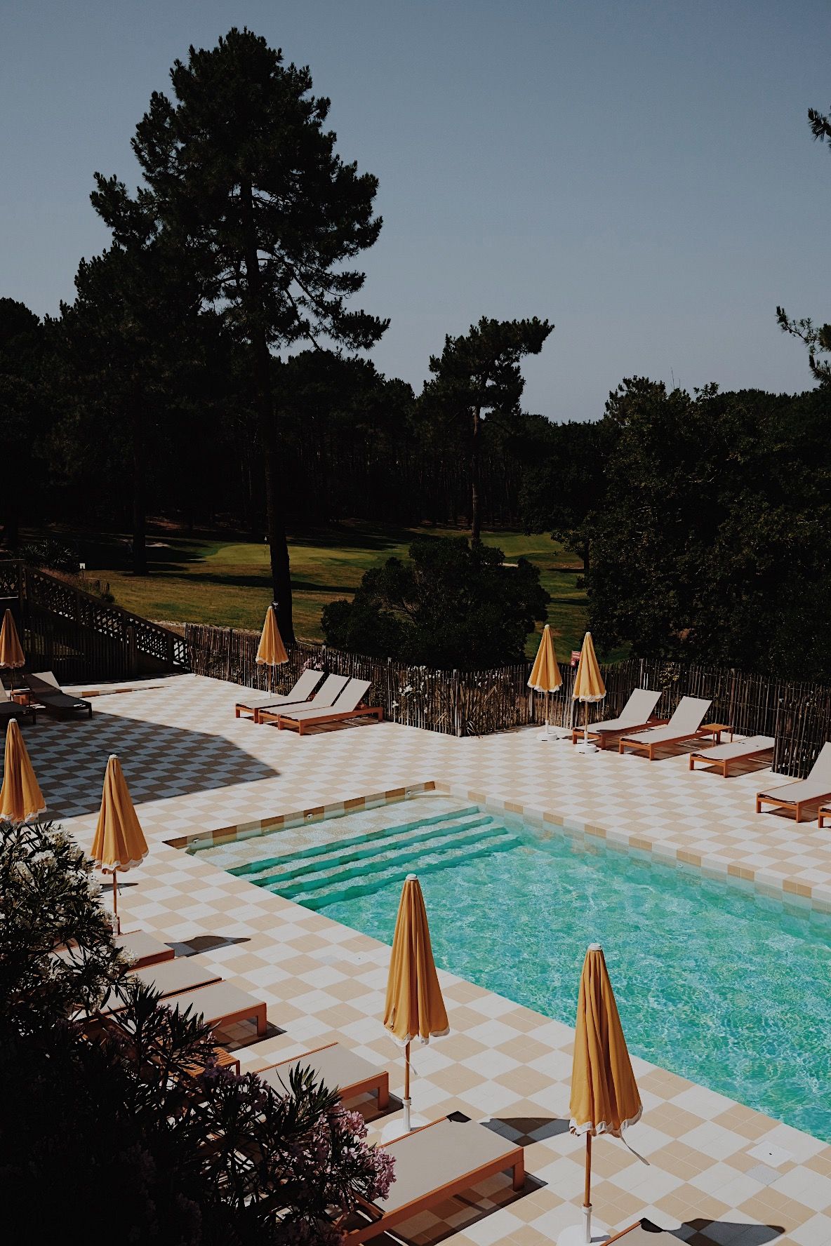 Zona de piscina con sillas de descanso y sombrillas, rodeada por una valla y árboles.