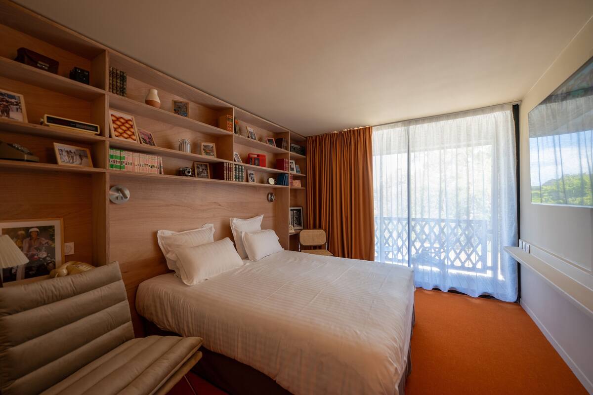 Habitación de hotel con una cama grande, una zona de estar con una silla, una estantería con diversos objetos y una ventana con cortinas.