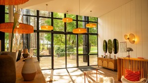 Un interior moderno con ventanas amplias, una mesa de madera con jarrones, un sillón blanco y mullido, y un tapete azul y blanco a rayas.