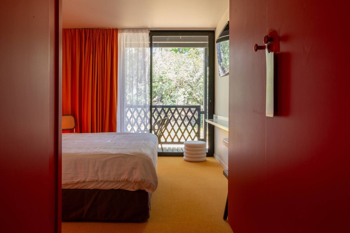 Una habitación de hotel con cama, ventana con cortinas, balcón con celosía y un pequeño taburete redondo.