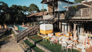 Un área de piscina con sillones de playa y sombrillas, una zona de estar cubierta con mesas y sillas, y un edificio con balcón y escaleras.