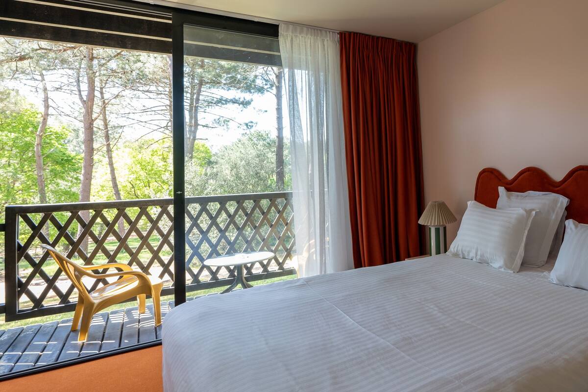 Habitación de hotel con una cama grande, ropa de cama blanca, vistas a árboles a través de una ventana, un balcón con una silla y un cabecero rojo.