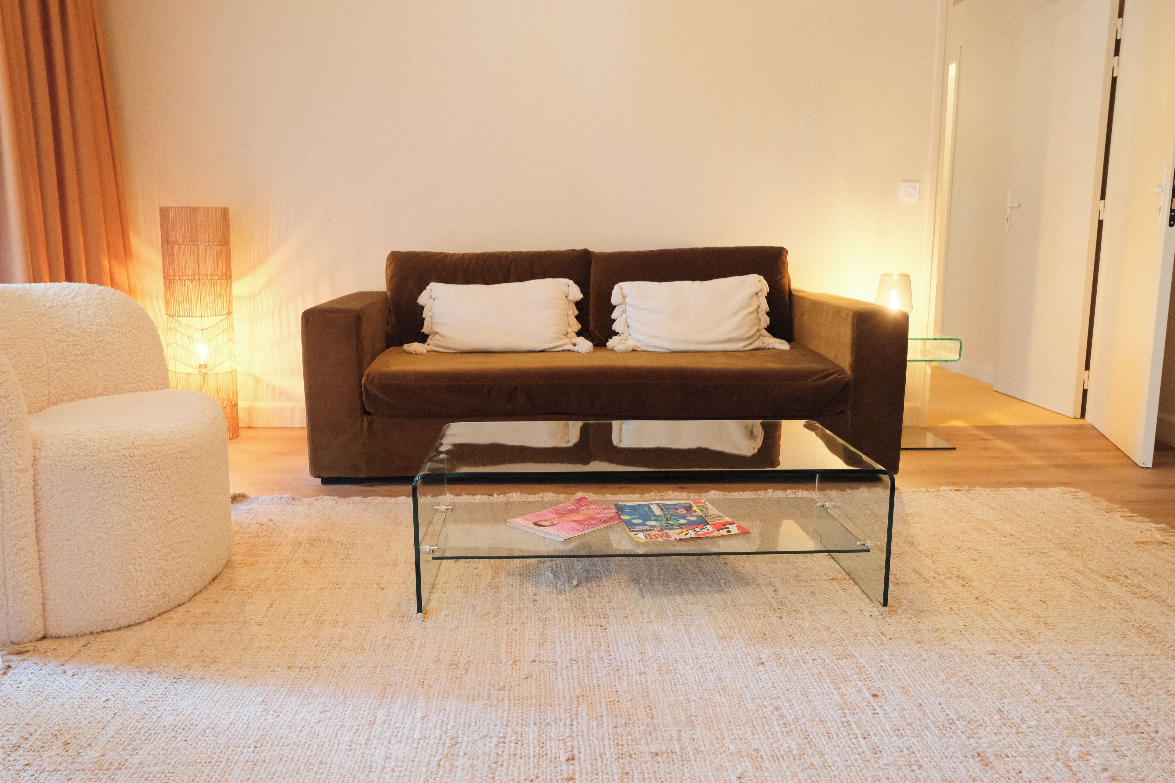 A living room with a brown sofa, two white cushions, a glass coffee table, and a white armchair.