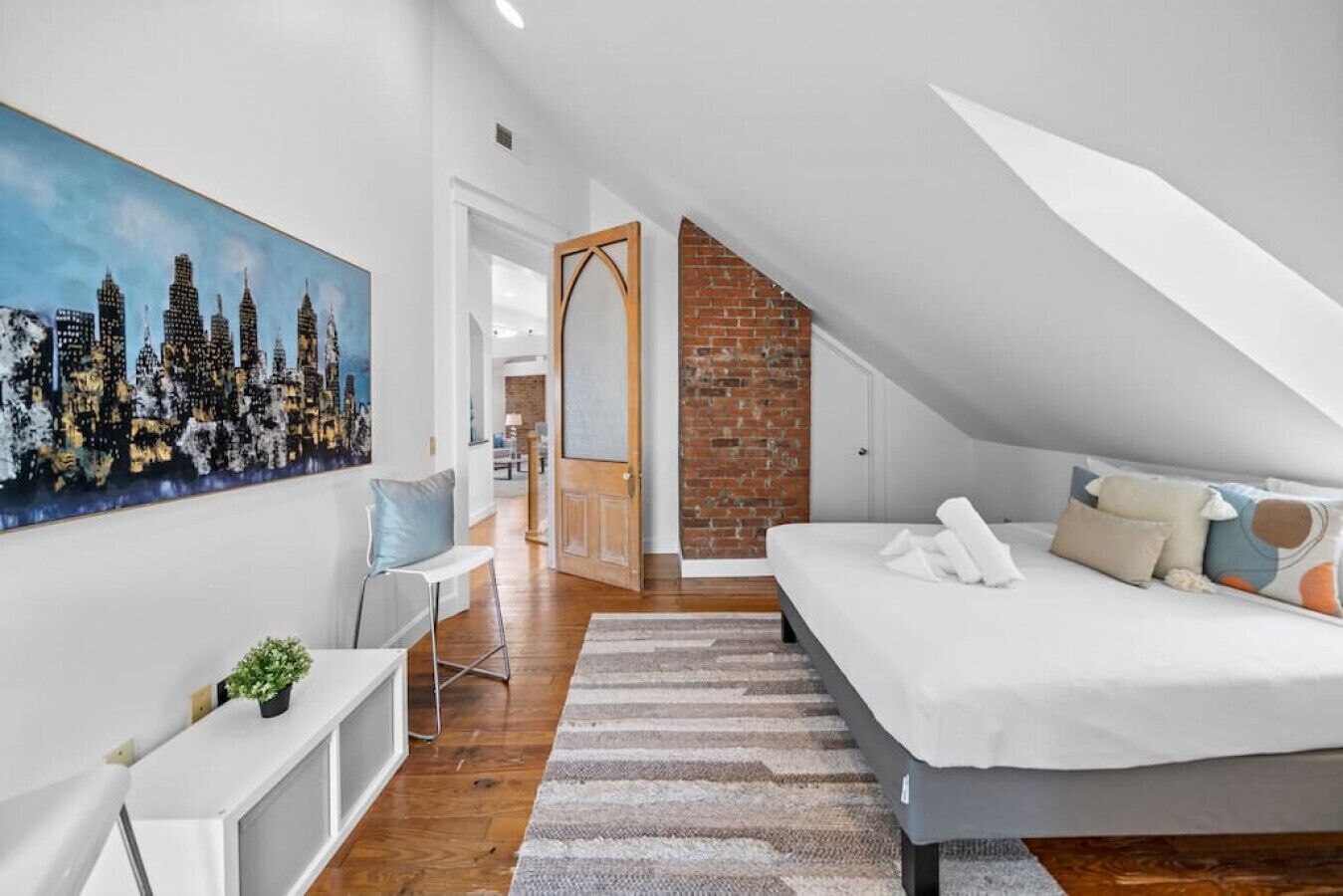 Une chambre moderne avec un plafond de style industriel, un tableau représentant un paysage urbain, un lit avec de nombreux coussins, un petit bureau et une chaise.