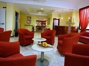 Un hall d’hôtel avec des fauteuils rouges, un comptoir d’accueil et, au fond, un espace réservé aux repas.