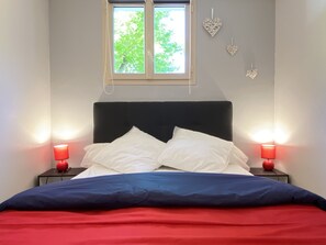 A bed with white pillows and a blue and red blanket, two bedside tables with red lamps, a window with a view of green trees, and two heart-shaped wall decorations.