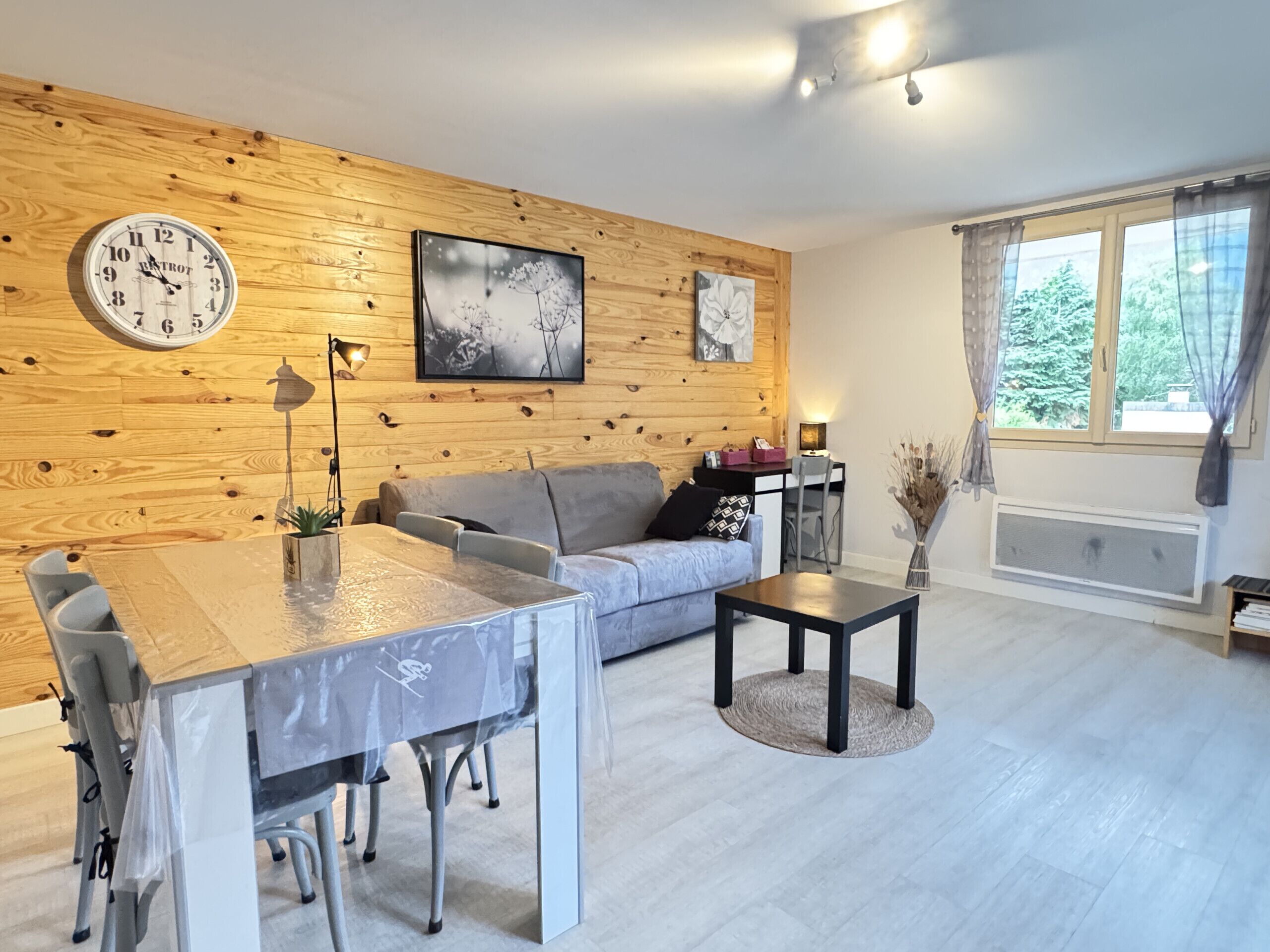 A modern living room with a wooden wall, a round clock, a sofa, a dining table, and a window with curtains.