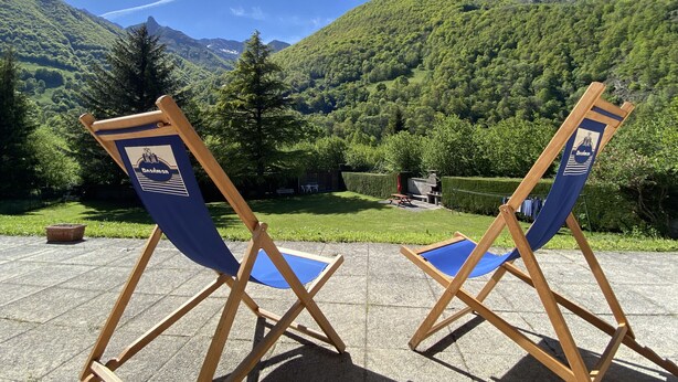 Zwei hölzerne Klappstühle mit blauen Kissen und einem Logo, auf befestigtem Untergrund mit Blick auf eine Berglandschaft.