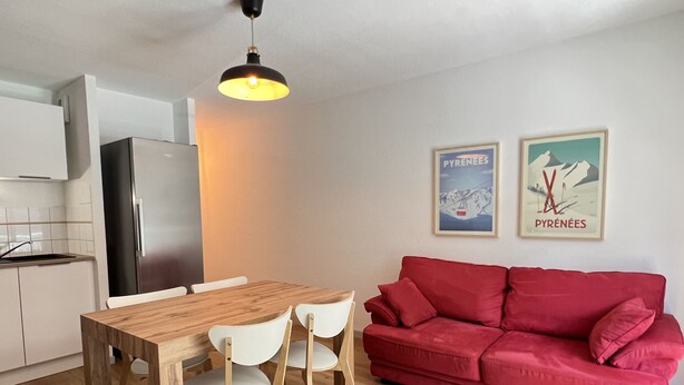 Una cocina moderna con armarios blancos, un refrigerador negro y una mesa de comedor de madera con sillas blancas. Un sofá rojo está apoyado en la pared, y hay dos cuadros enmarcados encima.
