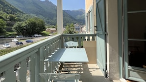 A balcony with a table and chairs, overlooking a road with parked cars and a mountain landscape. - T3 centre ville avec parking, balcon et wifi. 5P. (Cauterets)