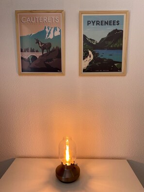Zwei gerahmte Poster mit Landschaftsansichten von 'Cauterets' und den 'Pyrenäen', sowie ein Tisch mit einer beleuchteten Glühbirnenlampe.