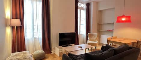Ein Wohnzimmer mit einer Couch, einem Fernseher, einer Lampe sowie einem Essbereich mit Tisch und Stühlen.