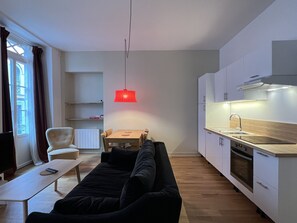 Een moderne keuken met witte kasten, een houten aanrechtblad en een ingebouwde oven. Er is een eettafel met stoelen en een rode hanglamp erboven. Een zwarte bank en een kleine witte stoel staan in de woonkamer, met een televisie op een dressoir.
