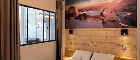 A hotel room with a bed, a window with curtains, a wall-mounted light fixture, and a painting of a mountainous landscape.