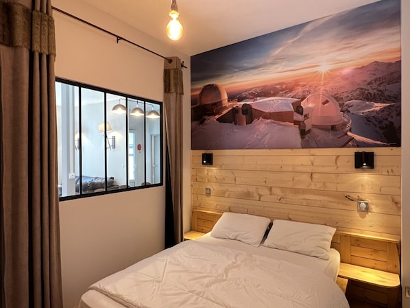 A hotel room with a bed, a window with curtains, a wall-mounted light fixture, and a painting of a mountainous landscape.